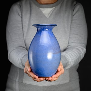 May include: A ceramic vase with a smooth, glossy finish in shades of vibrant blue. The vessel features a rounded, bulbous body that tapers upward into a narrow neck with a flared, circular rim. The glaze shows subtle variations in color intensity.