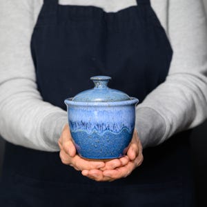 May include: A handmade, blue ceramic lidded jar with a speckled finish. The jar has a rounded shape with a lid and a small handle. The color transitions from a lighter blue at the top to a darker blue at the base, creating a textured appearance.