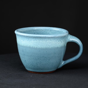 May include: A ceramic mug with a speckled light blue glaze that transitions to a paler, creamy blue near the rim. The mug features a rounded body, a curved handle on the right side, and a visible reddish-brown clay base at the bottom.