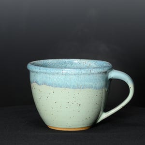 May include: A handmade ceramic mug with a light blue rim and handle, transitioning to a speckled, pale green body. The mug is filled with a hot beverage, indicated by the rising steam. The mug sits on a dark surface.