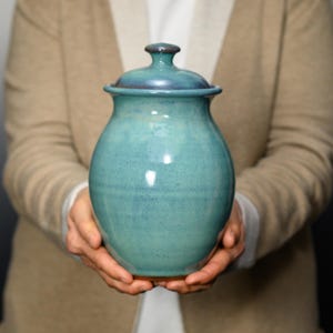 May include: A ceramic jar with a rounded body and a matching lid, featuring a glossy teal and blue glaze with subtle variations in color. The jar has a smooth, curved silhouette and a small knob handle on top of the lid, showcasing a handcrafted finish.