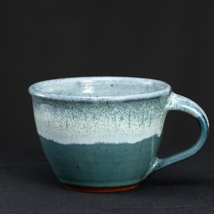 May include: A ceramic mug featuring a two-tone glaze with a dark teal base that transitions into a speckled white and light blue upper section. The mug has a wide, rounded bowl shape, a thick handle on the right side, and a visible terracotta-colored base.