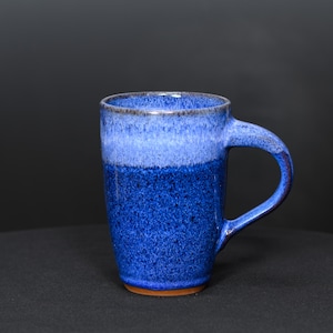 May include: A ceramic mug featuring a deep blue speckled glaze on the lower portion that transitions into a lighter, pale blue glaze near the rim. The mug has a sturdy, curved handle and a visible unglazed brown clay base at the bottom edge.