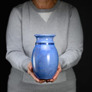 May include: A ceramic vase with a rounded body and a flared rim, featuring a variegated blue glaze with darker blue accents around the neck and a smooth, glossy finish that reflects light across its curved surface.