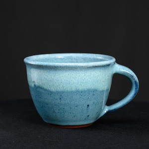 May include: A ceramic mug featuring a two-tone blue glaze with a lighter sky blue upper section and a darker, speckled blue lower section. The mug has a rounded body, a wide rim, and a curved handle, with a visible reddish-brown base.