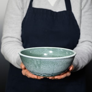 May include: A handmade ceramic bowl with a speckled, teal glaze. The bowl has a rounded shape with a slightly flared rim and a white edge. The bowl is held by a person wearing a gray sweater and a dark blue apron.