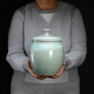 May include: A ceramic jar with a lid, featuring a light mint green glaze that transitions to a darker, speckled finish toward the base. The jar has a rounded, bulbous body and a small, knob-style handle on the center of the lid.