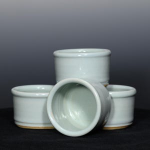 May include: Four small, light blue ceramic cups with a rounded shape. The cups are stacked and arranged, with one cup tilted to show the interior. The rims and bases are a slightly darker shade.