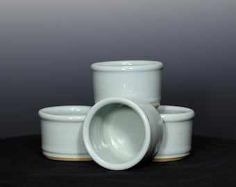 May include: Four small, light blue ceramic cups with a rounded shape. The cups are stacked and arranged, with one cup tilted to show the interior. The rims and bases are a slightly darker shade.