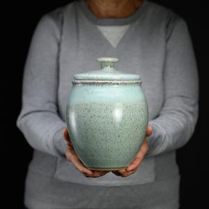 May include: A ceramic jar with a lid, featuring a light mint green glaze that transitions to a darker, speckled brown finish near the rim and base. The vessel has a rounded, bulbous body and a small, knob-like handle on the lid.
