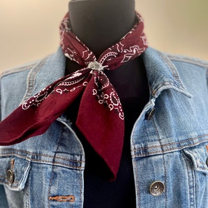 May include: A burgundy bandana with a white paisley pattern is secured with a silver-colored ring. The bandana is worn around a black mannequin neck, paired with a denim jacket. The jacket has silver buttons and a classic design.
