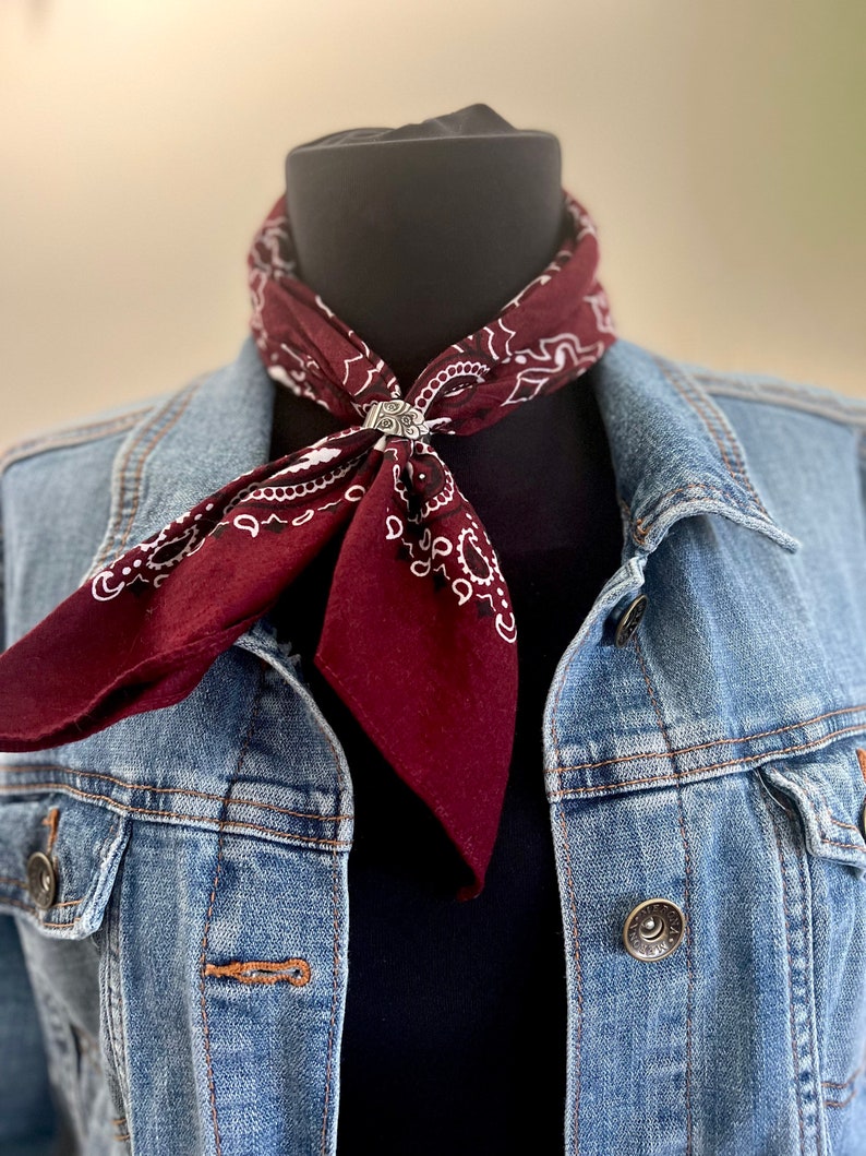 May include: A burgundy bandana with a white paisley pattern is secured with a silver-colored ring. The bandana is worn around a black mannequin neck, paired with a denim jacket. The jacket has silver buttons and a classic design.