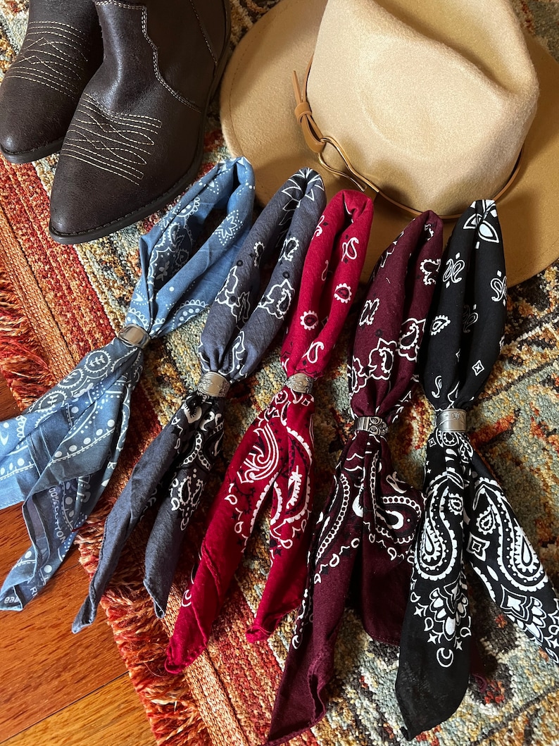 May include: An assortment of bandanas in various colors, including blue, red, and black, each secured with a silver ring. A brown cowboy hat and boots are also visible, suggesting a Western theme.