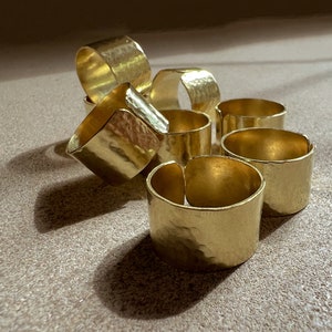 Op de afbeelding: Een verzameling goudkleurige servetringen met een gehamerde textuur. De ringen zijn cilindrisch en in een losse stapel gerangschikt, waarbij sommige elkaar overlappen. De belichting creëert highlights en schaduwen.