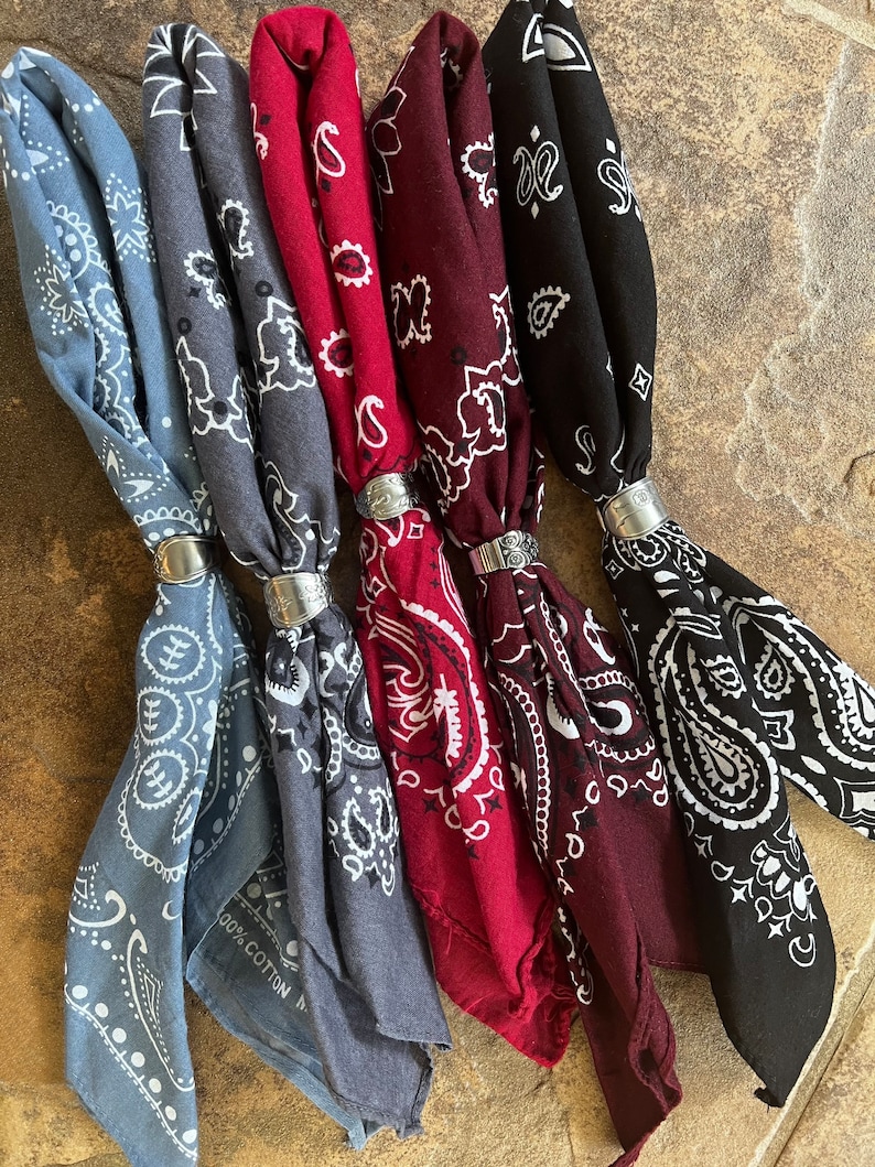 May include: A collection of six bandanas in various colors, including light blue, gray, burgundy, and black. Each bandana features a white paisley pattern and is held together with a silver-colored ring. The bandanas are made of 100% cotton.