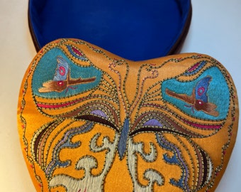 May include: Heart-shaped, open box with a mustard-yellow exterior and a blue interior. The lid is decorated with an embroidered butterfly design in shades of blue, purple, and gold. The box is likely used for jewelry or small keepsakes.