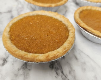 May include: Four individual mini pumpkin pies in aluminum pie tins on a white marble surface. The pies have a golden brown crust and a smooth, golden brown filling.