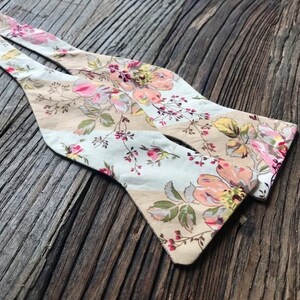 May include: A floral patterned bow tie with a light blue and beige background. The bow tie has a pink, yellow, and green floral pattern.