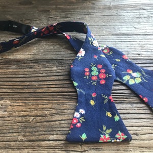 May include: A navy blue bow tie with a floral pattern featuring red, white, and green flowers. The bow tie is made of a soft, cotton fabric.