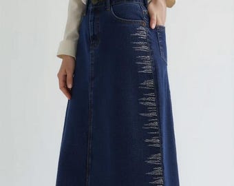 May include: A dark blue denim maxi skirt with a decorative white stitched design along the side. The skirt has a classic five-pocket design and a long, flowing silhouette. The model is wearing black high-heeled sandals.