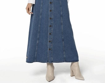 May include: A long denim skirt in a medium blue wash, with buttons down the front, worn with a white wrap-style blouse. The skirt has vertical seams. The model is wearing beige ankle boots.