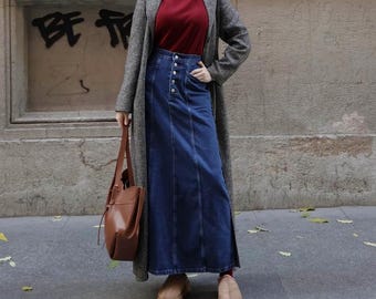 May include: A woman wearing a long, grey tweed coat over a red turtleneck sweater and a long, dark blue denim skirt with button detailing. She is carrying a brown leather tote bag and wearing brown leather clogs.