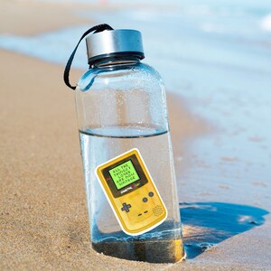 May include: A clear glass water bottle with a black lid and a yellow Game Boy Color sticker on the side. The sticker says "All the things I loved are now vintage".
