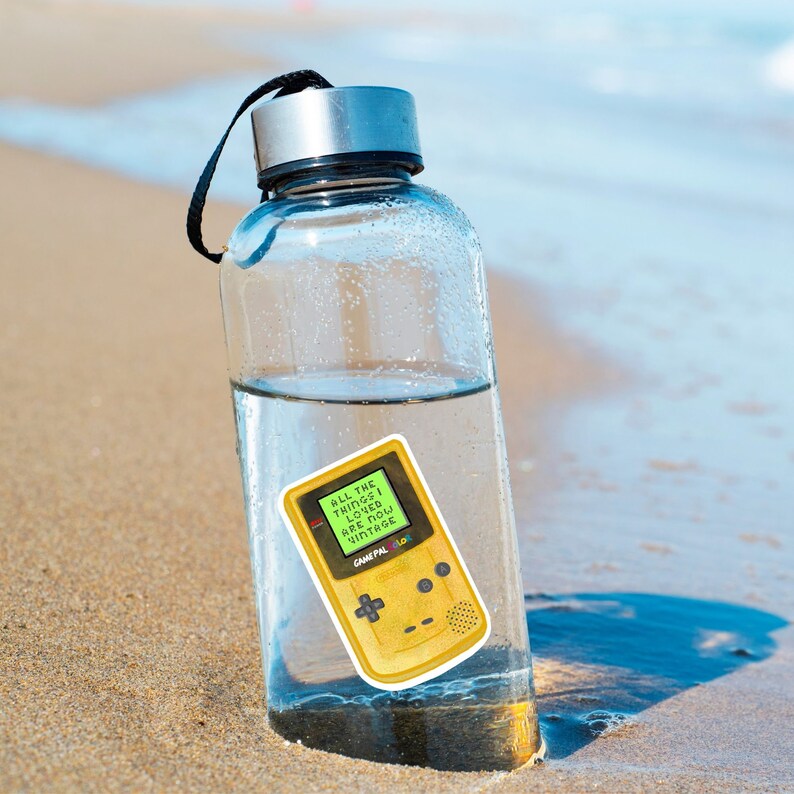 May include: A clear glass water bottle with a black lid and a yellow Game Boy Color sticker on the side. The sticker says "All the things I loved are now vintage".