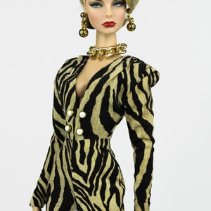 May include: A fashion doll wearing a black and tan zebra print jumpsuit with a gold chain necklace and earrings. The doll has blonde hair styled in a retro updo and red lipstick.
