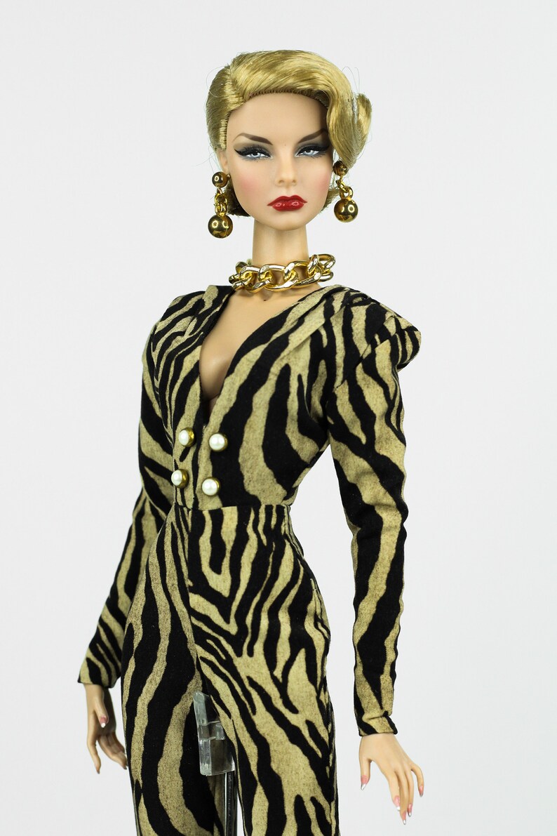 May include: A fashion doll wearing a black and tan zebra print jumpsuit with a gold chain necklace and earrings. The doll has blonde hair styled in a retro updo and red lipstick.