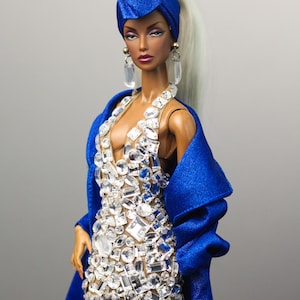 May include: A fashion doll wearing a royal blue turban, matching coat, and a sparkling crystal-embellished gown. The doll has long, pale blonde hair and dangling earrings. The dress features a plunging neckline and a high slit.