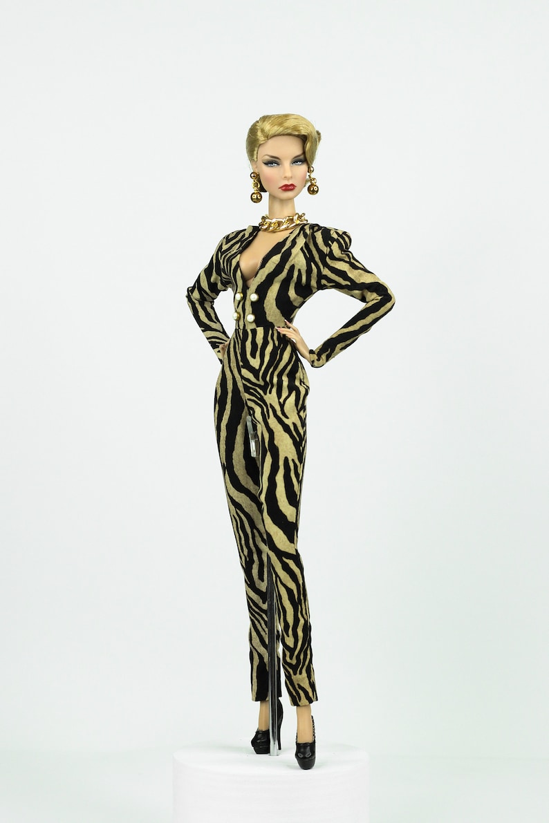 May include: A fashion doll wearing a black and gold zebra print jumpsuit with a plunging neckline, gold necklace, and gold hoop earrings. The doll is standing on a white platform.