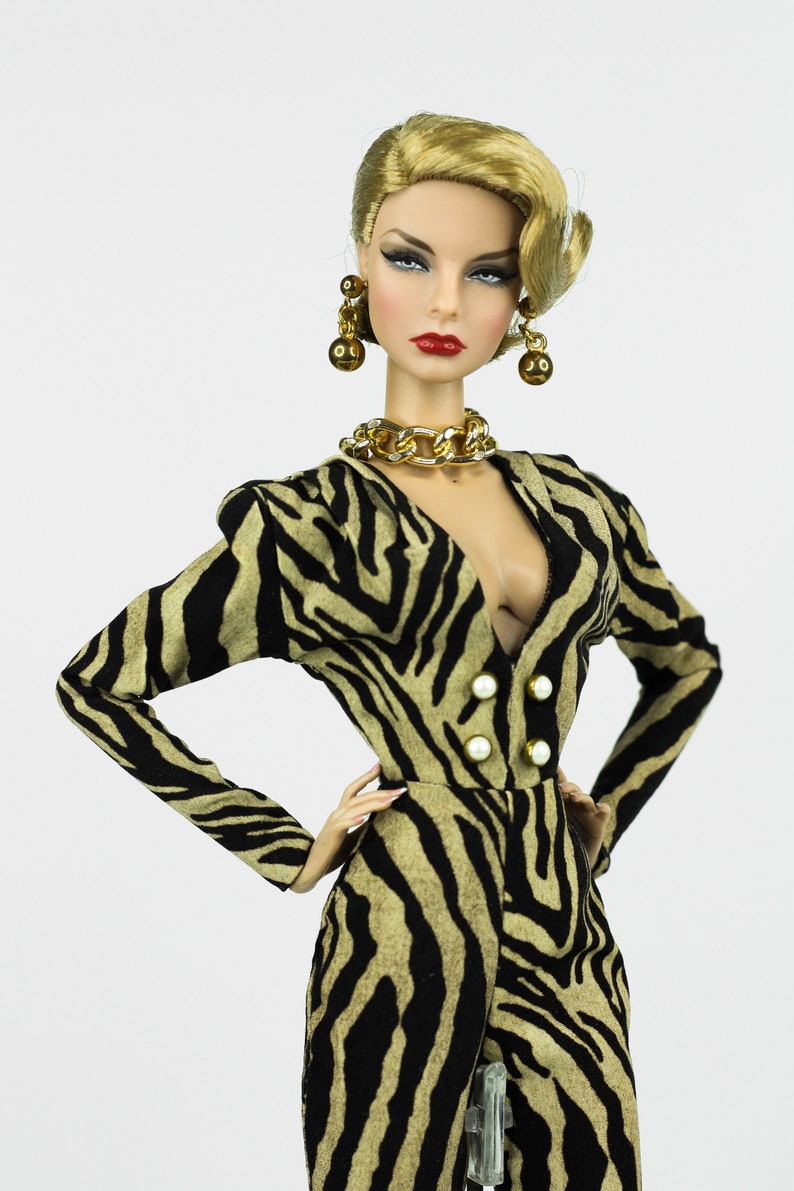 May include: A doll wearing a black and brown zebra print jumpsuit with a gold chain necklace and gold earrings. The doll has blonde hair styled in a wave and red lipstick.