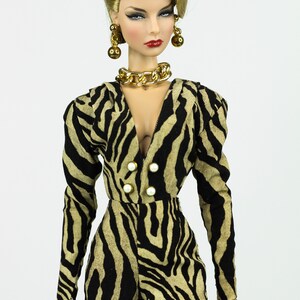 May include: A doll wearing a black and tan zebra print jumpsuit with a gold chain necklace and gold earrings. The doll has blonde hair and red lipstick.