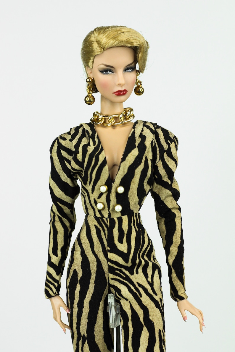 May include: A doll wearing a black and tan zebra print jumpsuit with a gold chain necklace and gold earrings. The doll has blonde hair and red lipstick.