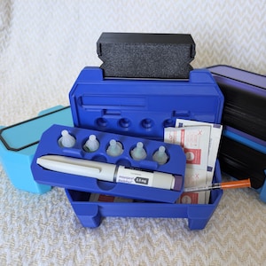 Puede incluir: Un conjunto de suministros médicos, que incluye un estuche azul con un bolígrafo blanco, agujas y toallitas con alcohol. La imagen también muestra un estuche azul y otro morado, y un bloque de espuma negro.