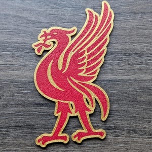 Puede incluir: Un emblema rojo y dorado del Liver Bird, una criatura mítica, se representa sobre un fondo gris. El ave tiene las alas extendidas y está de pie. El emblema es un símbolo de la ciudad de Liverpool.