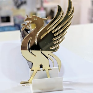 May include: A gold-coloured, stylised bird emblem with outstretched wings, holding a sprig in its beak. The emblem is mounted on a white base. The bird's design features a sleek, modern aesthetic, with a reflective surface.