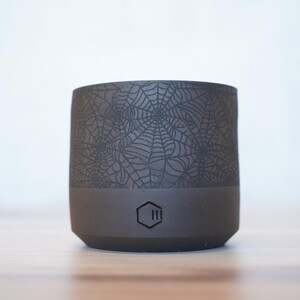 May include: A dark gray ceramic tumbler with a spiderweb design on the upper portion. The bottom section is a solid dark gray, and a geometric logo is visible. The tumbler is cylindrical and sits on a wooden surface.