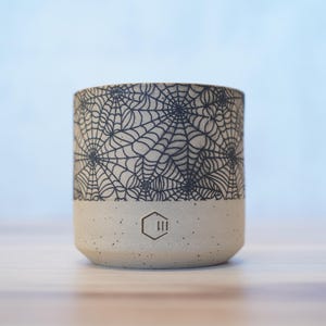 Könnte beinhalten: Ein zylindrischer Keramikbecher mit beigefarbenem Boden und einem schwarzen Spinnennetz-Design auf dem oberen Teil. Der Becher hat eine gesprenkelte Textur und ein geometrisches Logo in der Nähe des Bodens. Der Becher wird wahrscheinlich für Getränke verwendet.
