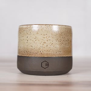 May include: A cylindrical ceramic mug featuring a two-tone design with a speckled cream-colored upper section and a solid dark charcoal-colored base. A small black hexagonal logo with three vertical lines inside is centered on the dark base.