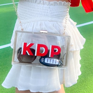 May include: A clear acrylic box clutch bag with a gold chain strap. The bag has the letters "KDP" in red letters on the front. The bag also contains a car key and a pair of sunglasses.