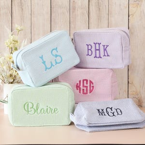 May include: Five rectangular seersucker cosmetic bags in light blue, lavender, pink, mint green, and blue-and-white stripe. Each bag features custom embroidery, including monograms LS, BHK, KSG, MGD, and the name Blaire in various thread colors.
