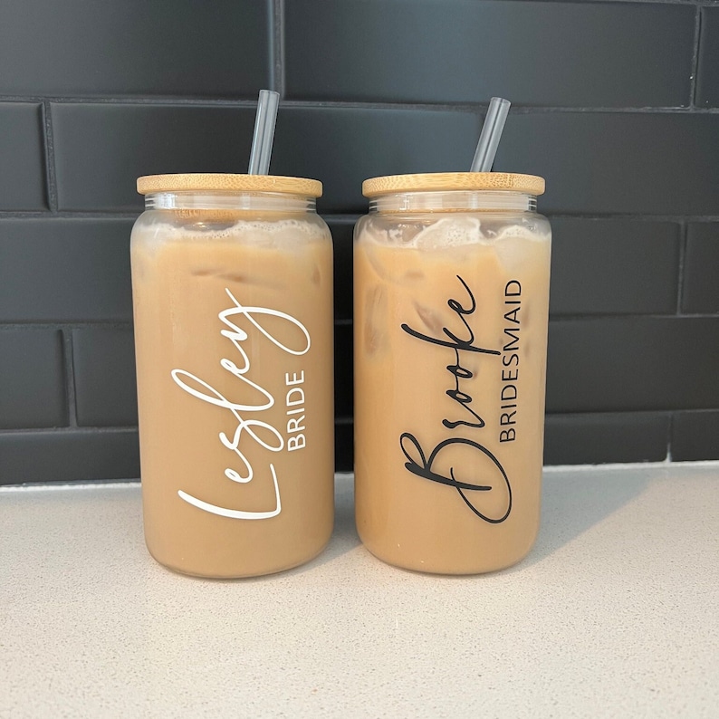 May include: Two clear glass tumblers with bamboo lids and straws. The tumblers are filled with a light brown beverage. One tumbler has the name "Lesley" written in white with the word "Bride" underneath. The other tumbler has the name "Brooke" written in black with the word "Bridesmaid" underneath.