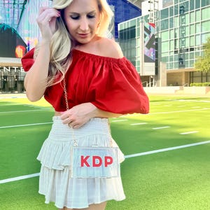 Pode incluir: Uma mulher vestindo um top vermelho de ombro ca&iacute;do e uma saia branca com babados. Ela est&aacute; carregando uma bolsa de m&atilde;o transparente de pl&aacute;stico com as letras "KDP" em vermelho.