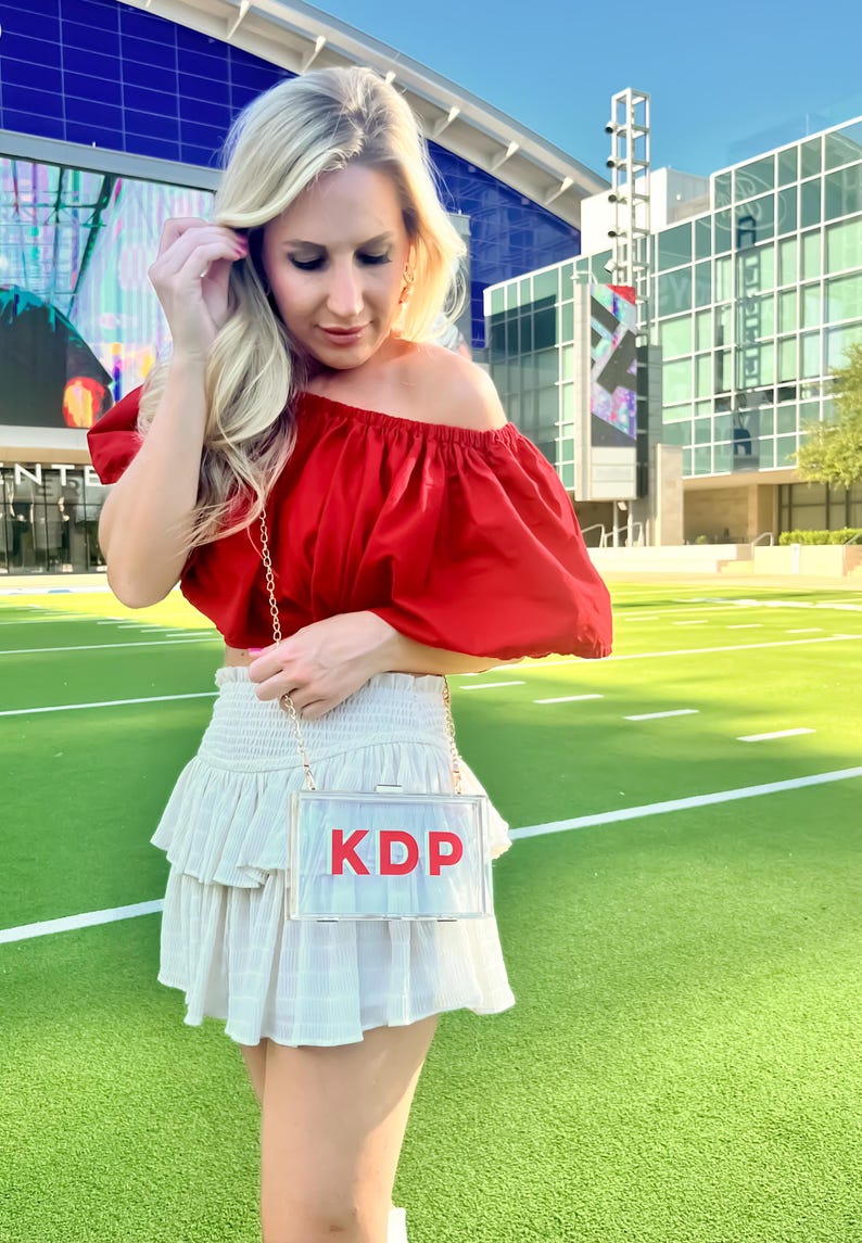 Pode incluir: Uma mulher vestindo um top vermelho de ombro ca&iacute;do e uma saia branca com babados. Ela est&aacute; carregando uma bolsa de m&atilde;o transparente de pl&aacute;stico com as letras "KDP" em vermelho.