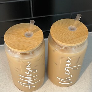 May include: Two clear glass tumblers with bamboo lids and clear straws. The tumblers are filled with iced coffee and have the names "Addison" and "Meagan" written on them in white.