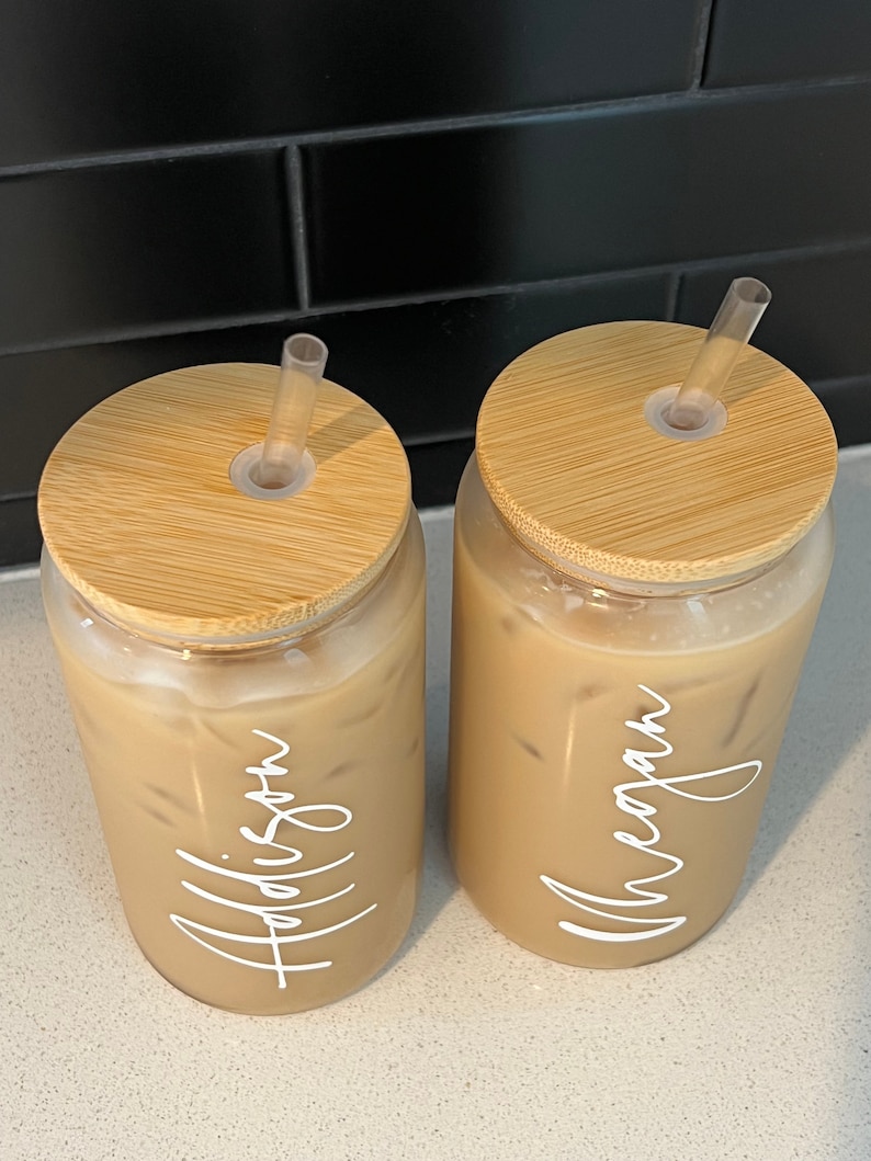 May include: Two clear glass tumblers with bamboo lids and clear straws. The tumblers are filled with iced coffee and have the names "Addison" and "Meagan" written on them in white.