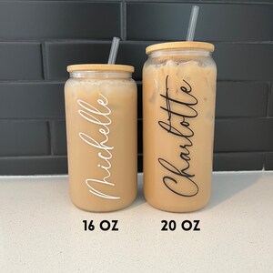 May include: Two clear glass tumblers with bamboo lids and straws. The tumblers are filled with a light brown liquid. The tumbler on the left is labelled "Michelle" and "16 OZ". The tumbler on the right is labelled "Charlotte" and "20 OZ".