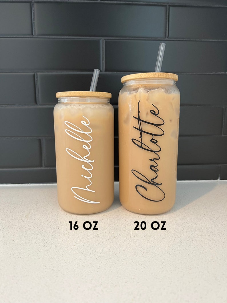 May include: Two clear glass tumblers with bamboo lids and straws. The tumblers are filled with a light brown liquid. The tumbler on the left is labelled "Michelle" and "16 OZ". The tumbler on the right is labelled "Charlotte" and "20 OZ".